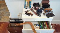 Overview showing sunglasses in cases, wristwatches, hunting cutting tool, dictionaries, briefcases, maps