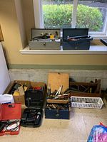 Overview of the lot showing several toolboxes and cardboard boxes filled with assorted hand and power tools against a wall below a window.