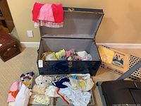 Open black trunk filled with various children's clothes, dolls, and a wool blanket with additional items laid out in front.