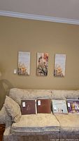 Three wall art panels mounted on a beige living room wall above a patterned couch. Each canvas is vertical and spaced evenly, each approximately 12x24 inches in size, depicting floral and natural themes.