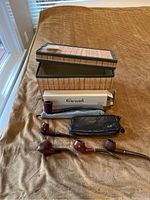 Six wood smoking pipes and various accessories displayed on a brown cloth with woven box backdrop.