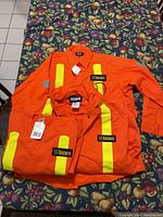 Three pieces of high-visibility orange construction safety wear showing City of Toronto patches and yellow vertical reflective stripes, arranged on a table with patterned cloth underneath.