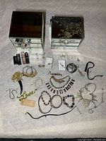 Photo showing two mirrored jewellery boxes, assorted jewelry including earrings, necklaces, bracelets, rings, and four lipsticks arranged on white surface.