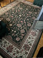 Photo showing the full rug laid out on a hardwood floor with a floral design and contrasting border in cream and pink tones on a forest green background.