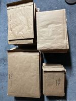 Four piles of brown padded shipping envelopes of varying sizes. Larger mailers on the left, smaller on the bottom right, all rectangular with adhesive flaps.