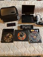 Photo showing the Toshiba DVD player with fold-up screen, remote control, power cable, rechargeable battery, black travel case, and three DVDs in sleeves labeled Pulp Fiction, Any Given Sunday, Wild Boys Season 2.