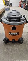 Front view showing orange tank with Ridgid logo, power intake on the front and four caster wheels.