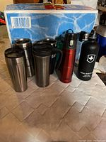 Eight travel mugs and bottles arranged on floor, brands visible include Starbucks, Sigg, Thermos.