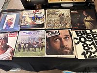 Photo showing seven comedy LP records on a black tablecloth, featuring covers of LBJ Back at the Ranch, George Carlin, Bill Cosby, Rappin' Rodney, Monty Hall, and two others with illustrated covers.