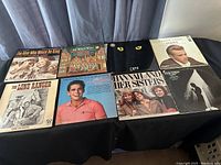 Shows a selection of 8 LP records laid flat on a black surface with a grey curtain backdrop. Titles visible include The Man Who Would Be King, The Lone Ranger, James Dean Giant, Dustin Hoffman Lenny, and others.