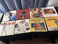 Eight LP soundtrack records arranged on black surface, showing titles like 'MASH', 'Outlaw Blues', 'Circus World', 'Easy Rider', 'W.C. Fields and Me', and 'A Man and a Woman'.