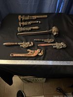 Nine vintage pipe wrenches and one large ratchet wrench arranged on a black cloth with a measuring ruler showing scale. Tools vary in size and style, with evident rust and wear.