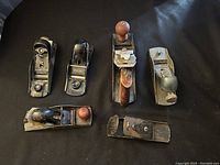Lot of six vintage carpenter hand planes including Stanley metal block planes and wood-metal combination planes, laid out on black surface.