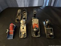 Six vintage metal hand planes arranged in two rows on black fabric background, showing size and style variety