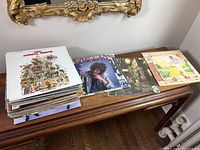 Stack of more than 20 vinyl albums on a wooden bench, mostly with visible spines and top covers. Assorted genres.