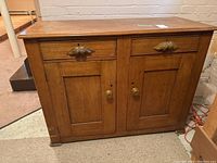 Full front view of wooden antique cabinet with two drawers and two doors, showing carved leaf-shaped handles and round knobs.
