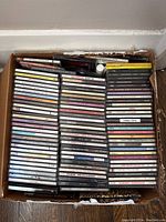 Top down view of a box full of assorted pop and rock music CD albums, showing multiple rows of jewel cases.