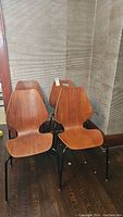 Full view of four teak veneer chairs with black metal legs against wall.