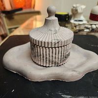 Photo of handmade jesmonite round decorative box with vertical ridges and lid with rounded knob, placed on an irregular wavy edged tray with gray marbled finish.