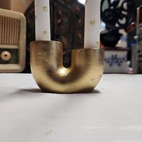 Close-up image of the gold-colored jesmonite candle holder shaped like a 'U', holding two white candles with gold heart patterns.