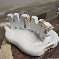 Close-up photo showing white jesmonite tray with block letters "HOME" and gold leaf accents on letter edges and tray corner.