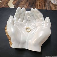 Front view of the white Jesmonite cupped hands ring holder with a gold ring inside and golden crystal decoration along one side.