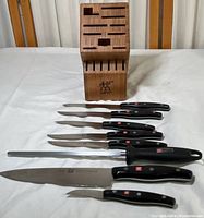 Knife set laid out with the wooden block, showing 7 knives and a sharpening steel, all with black handles and J.A. Henckels logo visible on some handles and blades.