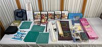 Overview of all needlework and crafting items arranged on a table showing DVDs, books, cutting mats, needles, threads, trays, tapes, and other crafting supplies