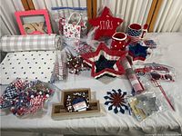 Photo showing various patriotic themed crafting and decoration items arranged on a table including star-shaped dishes, fabric rolls, mugs, wooden letters, and small embellishments.