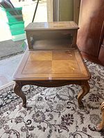 Photo of an antique two tier wooden side table showing the tabletop and the smaller upper tier, placed on a patterned carpet indoors next to furniture pieces.