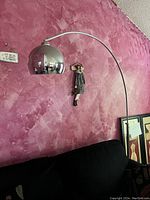 Full view of the chrome goose neck floor lamp mounted next to a pink textured wall and a black couch, showing the dome shade and curved arm.