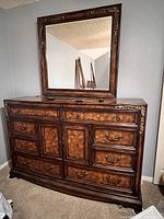 Front view of large wooden dresser with multiple drawers, cabinet doors, and matching mirror. Decorative brass-tone detail present on corners, drawers, and frame of mirror.
