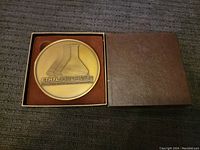 Top view of bronze medallion inside brown presentation box showing the medallion's raised design and company name.