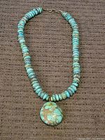 Full view of turquoise necklace showing round flat disk beads and pendant on woven brown fabric.