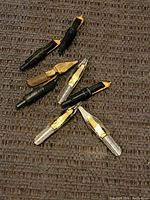 Photo showing seven fountain pen nib and feed units arranged on dark fabric, showing a mix of black plastic feeds and metallic nib sections in gold and silver tones.