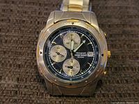 Close-up of Seiko men's chronograph watch face showing black dial, three sub-dials, tachymeter scale, date window, and gold-tone accents on bezel and crown.