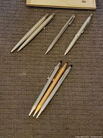Overview of seven pens on a textured cloth surface showing two Parker pens at top left, Sheaffer and Pentel pens to the right, and three Cross pens below in gold and silver finishes.