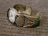 Side angle of the sterling silver cuff watch band with visible turquoise and coral chip inlay Thunderbird design and round watch face.