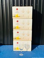 Four stacked boxes each showing two wine glasses and a yellow flower on the packaging. One wine glass placed in front for scale.