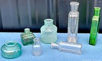 Seven antique glass bottles displayed on a blue cloth surface including green, clear, and aqua colors with various shapes