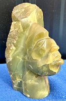 Side profile photo of hand-carved green onyx Aztec head showing translucent stone and carved facial details against black and blue background.