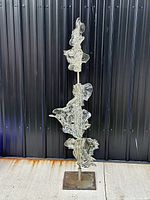 Full view of the vertical art glass sculpture with all four glass pieces mounted on a steel pole, placed outdoors against a dark vertical panel background.