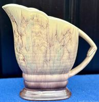 Side angle of beige to pale purple drip glaze jug showing scalloped top and curved handle.