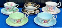 Five vintage fine bone china tea cups with matching saucers shown on white wood surface, colors include pastel green, blue, black with gold and white with pink floral patterns