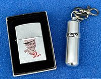 Zippo lighter in original black box next to silver metal Zippo lighter fluid keychain dispenser on blue cloth background.