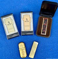 Six vintage refillable lighters displayed on blue cloth: two boxed Paris-Dakar cream-colored lighters, one lighter with tortoise shell inlay in a black case, and three loose metal lighters in various finishes and styles.