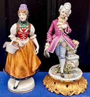 Two porcelain figurines side by side on a black background, showing the German Schwarzwalden figure in colorful traditional dress and the Italian Capodimonte figure in historical costume on a gold-toned base.