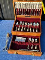 Flatware set displayed in an open wooden chest showing knives, forks, spoons, and serving tongs, arranged on maroon velvet lining.