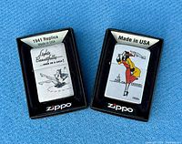Two Zippo lighters in original black display boxes shown on blue fabric surface. One lighter depicts 1941 replica design in brushed chrome, the other a colorful 1930's pin-up girl illustration.