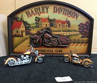 Wooden wall hanging with Harley Davidson logo and rural scene with houses, showing vintage motorcycle relief and two metal motorcycle figurines in front on black surface.